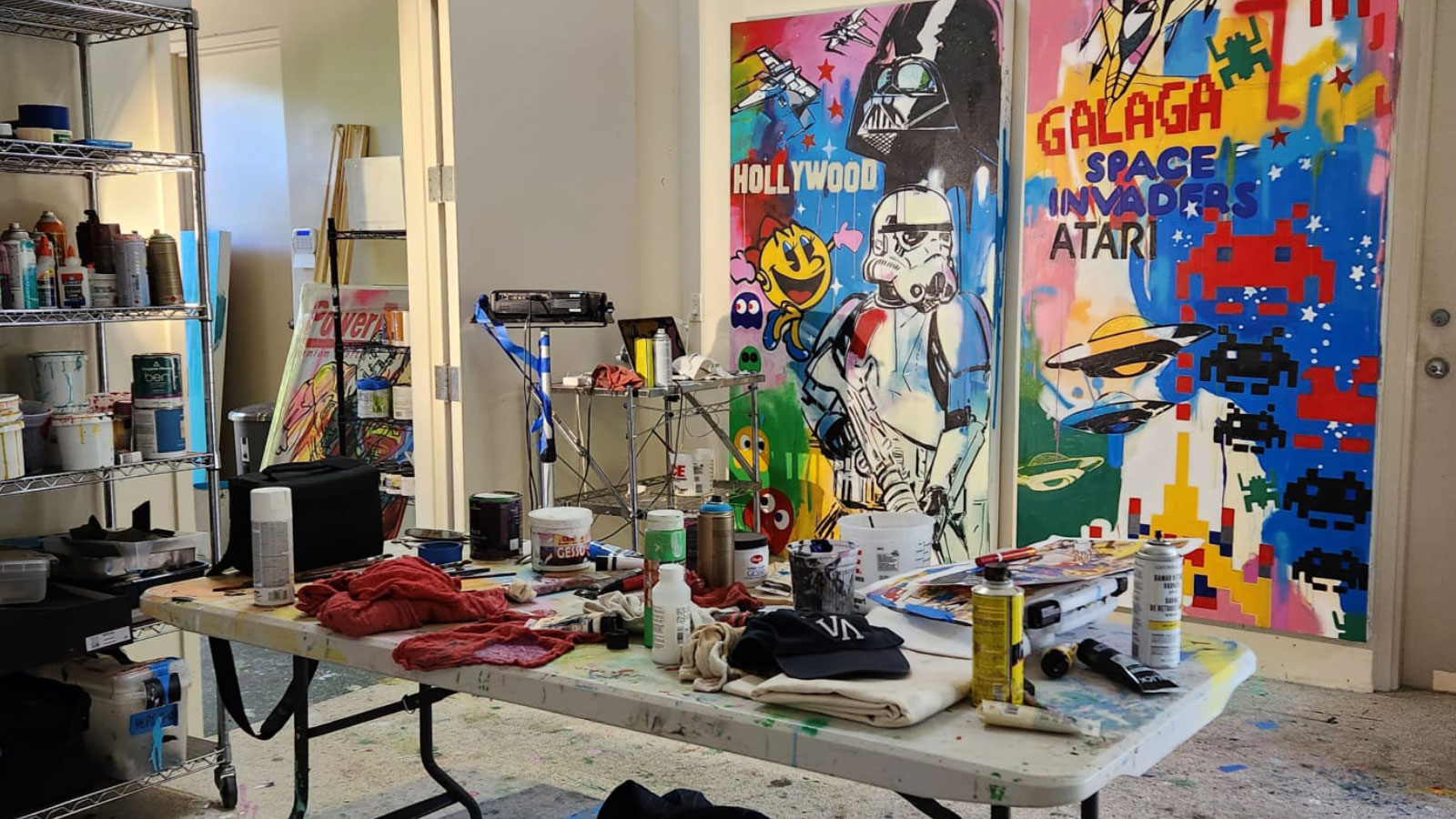Art painting studio with a table splattered with various colors of paint, assorted used liquid and spray paint cans, painting tools, shelves with paints, and a Star Wars and Space Invaders themed painting.